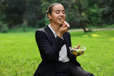 Lunch time. Happy businesswoman eating salad on green grass in park