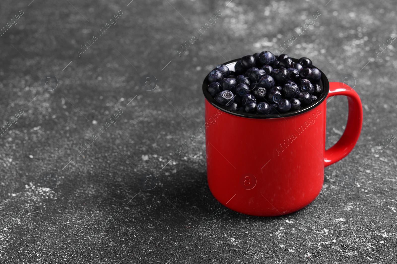 Photo of Ripe bilberries in mug on grey textured table. Space for text