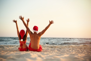 Photo of Lovely couple with Santa hats having fun together on beach, back view. Christmas vacation