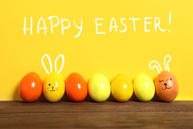 Two eggs with drawn faces and ears as Easter bunnies among others on wooden table against yellow background