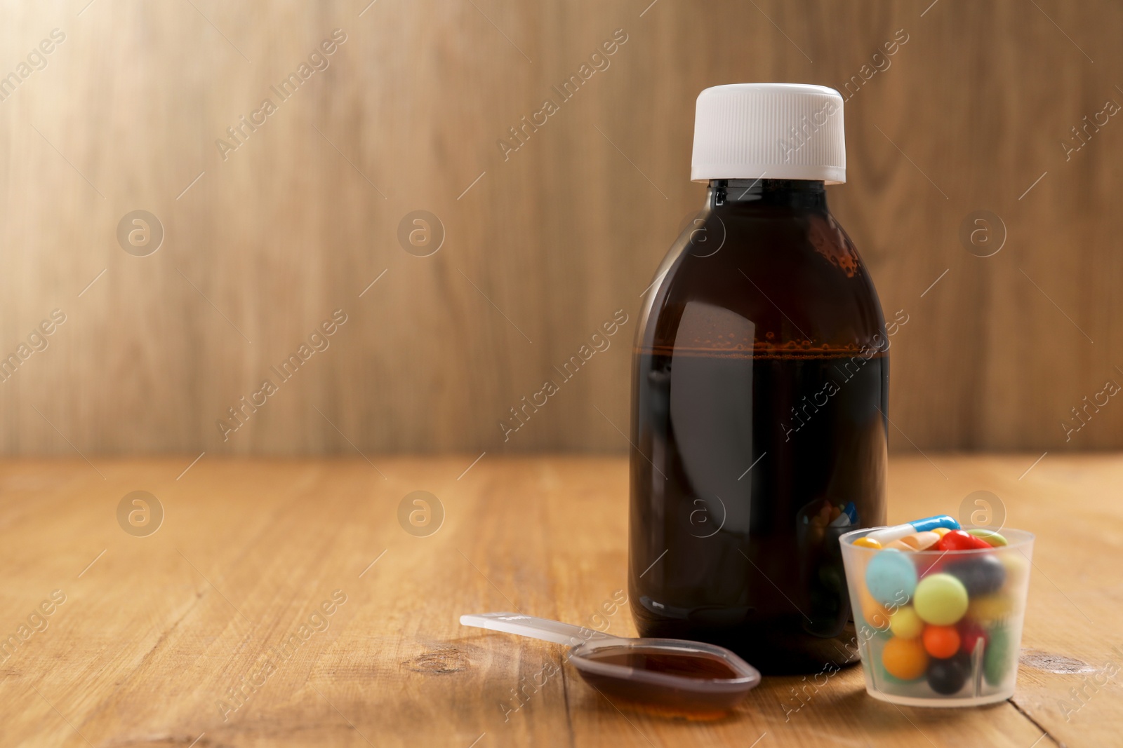 Photo of Bottle of syrup, dosing spoon and measuring cup with pills on wooden table, space for text. Cold medicine