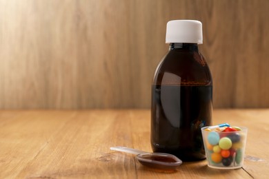 Bottle of syrup, dosing spoon and measuring cup with pills on wooden table, space for text. Cold medicine