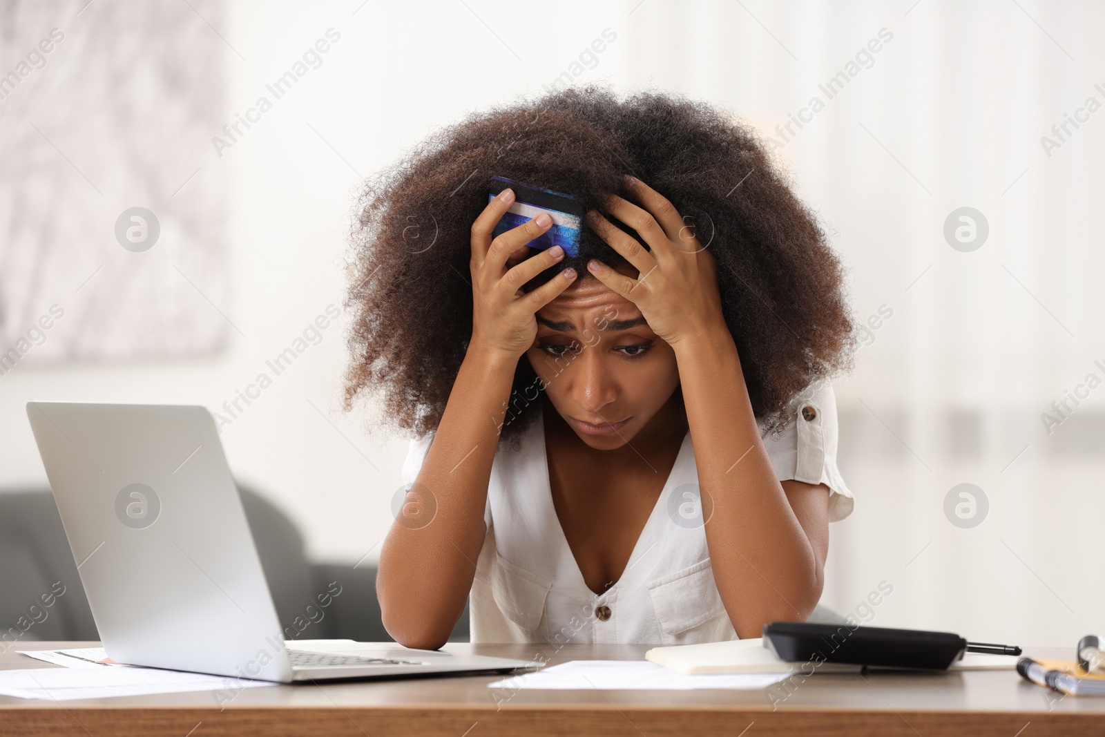 Photo of Upset woman with credit card and laptop at table indoors. Debt problem