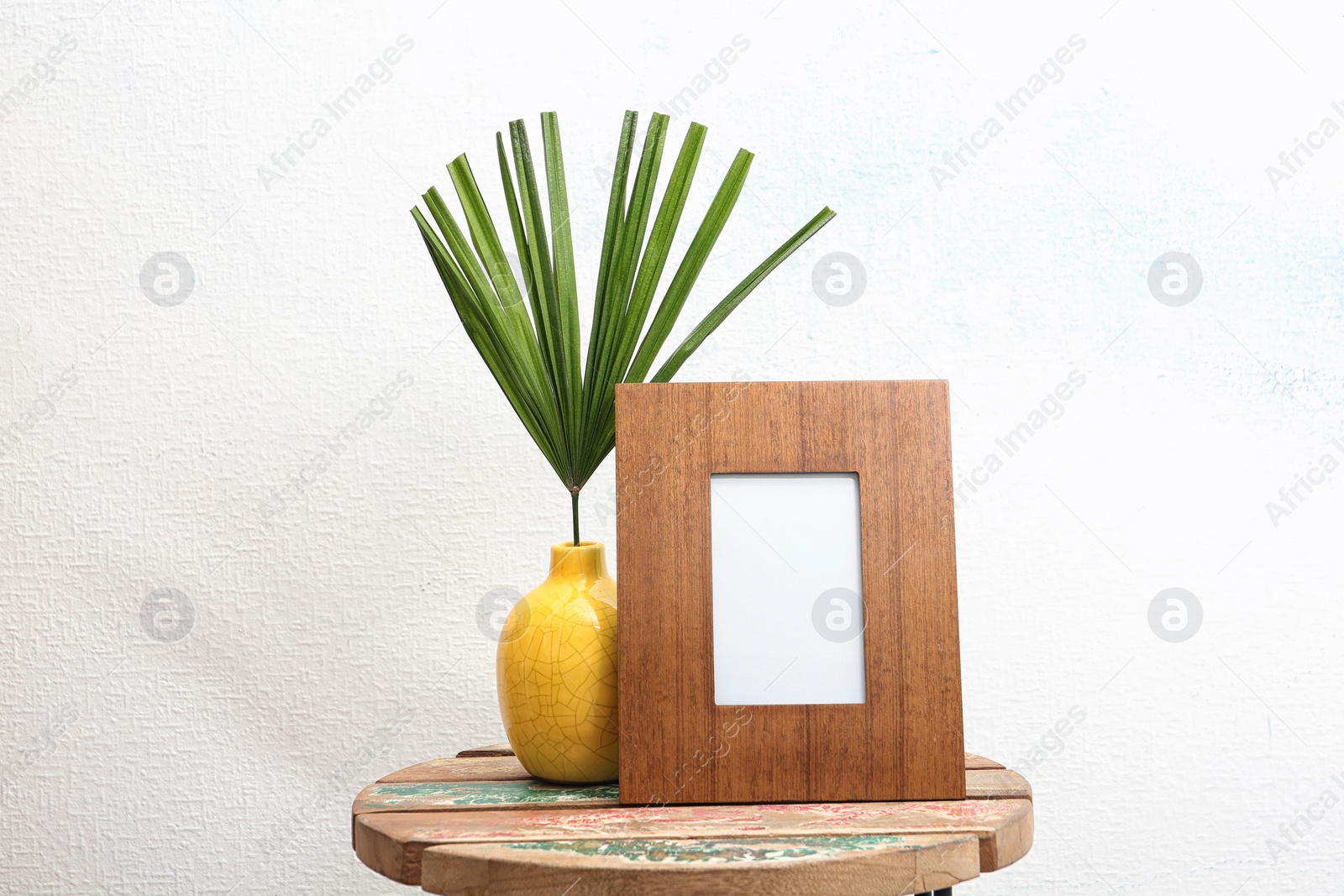 Photo of Blank frame and vase with tropical leaf on table near white wall. Mock up for design