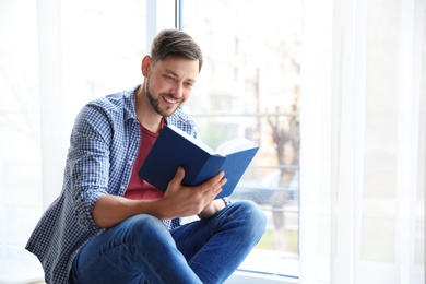 Handsome cheerful man reading book near window indoors. Space for text