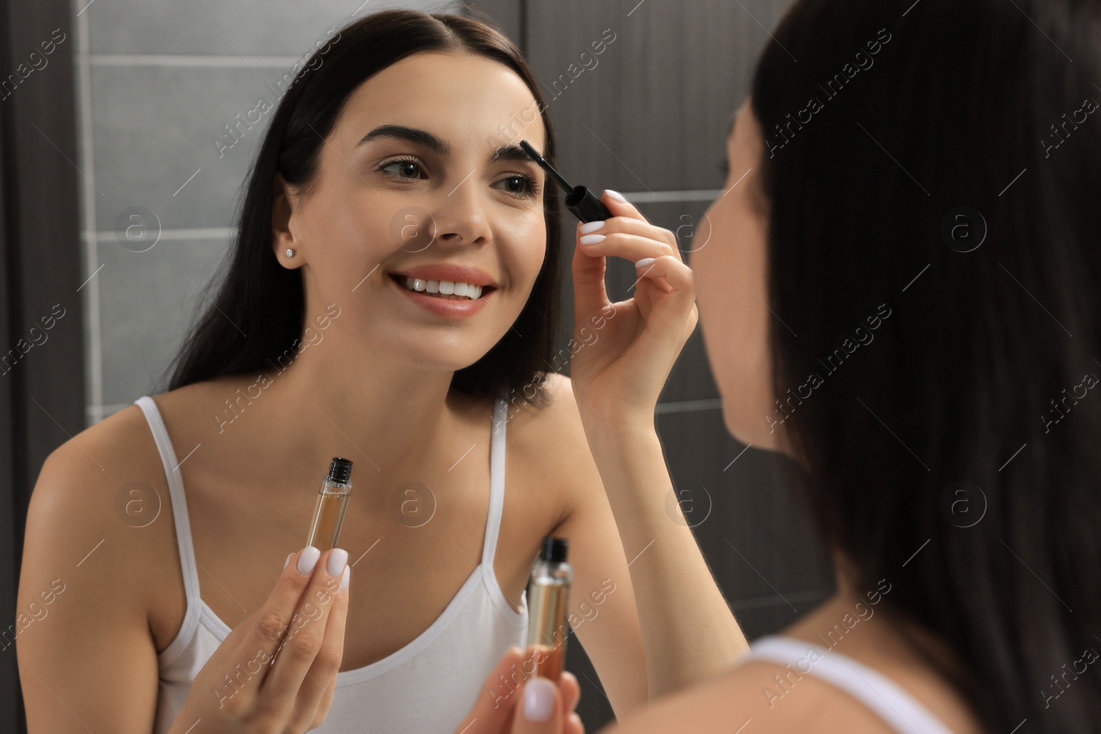 Photo of Young woman applying oil onto eyelashes near mirror indoors