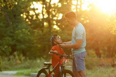 Photo of Dad putting bicycle helmet on son in park on sunny day