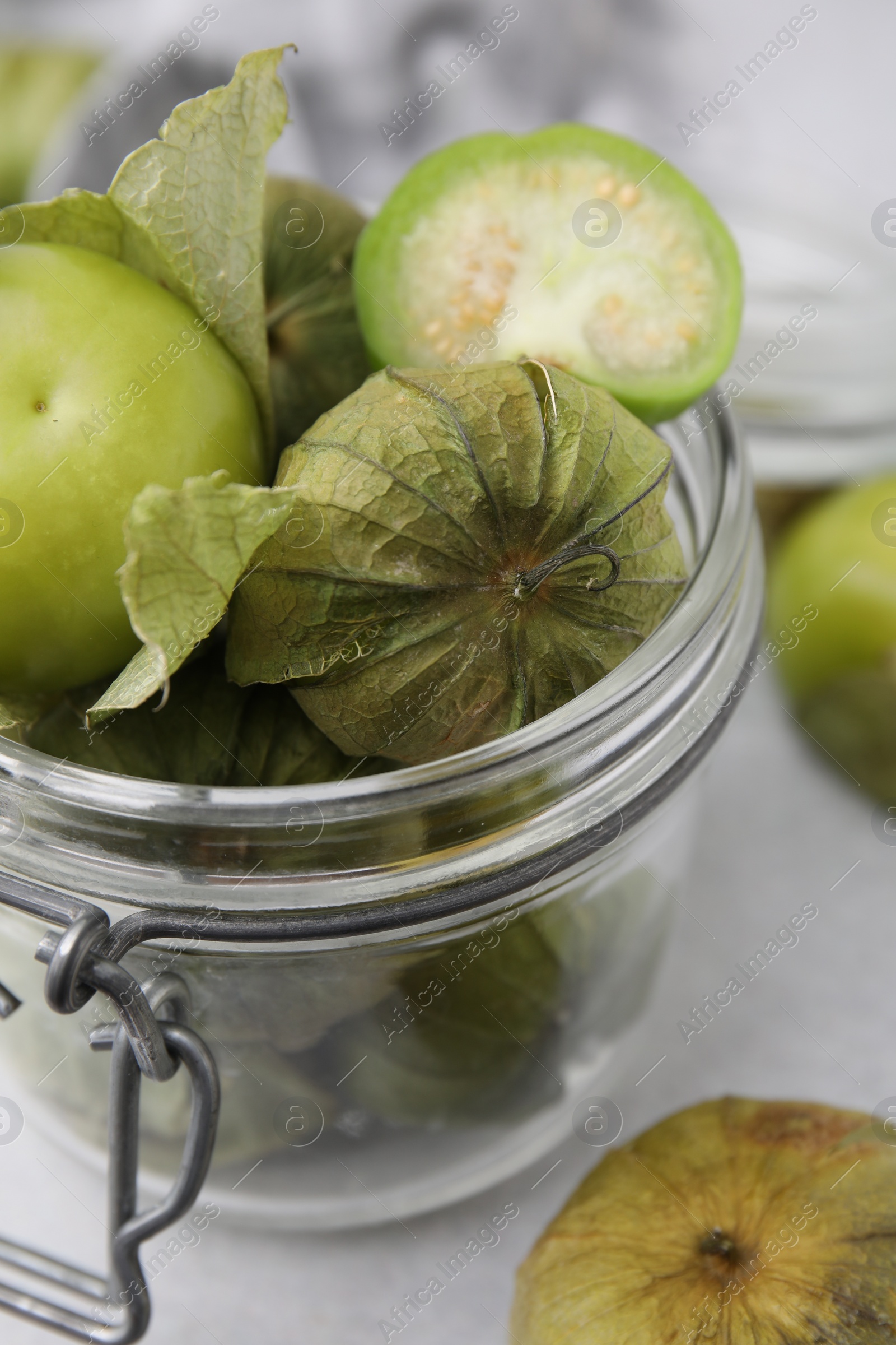 Photo of Fresh green tomatillos with husk in glass jar on light table, closeup