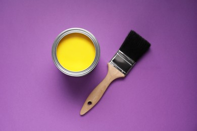 Photo of Can with yellow paint and brush on purple background, flat lay