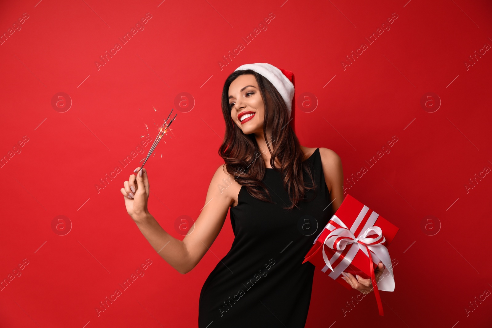 Photo of Beautiful woman wearing Santa hat with Christmas gift and bengal lights on red background