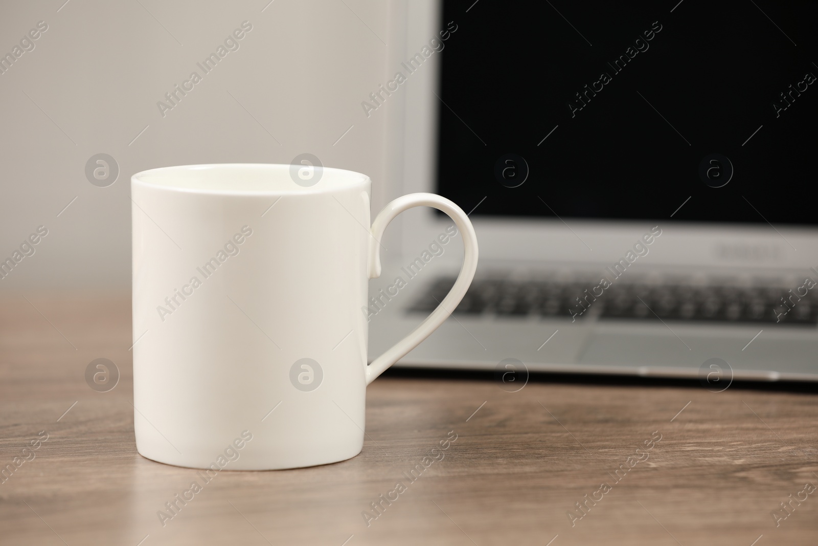 Photo of White ceramic mug and laptop on wooden table at workplace. Space for text