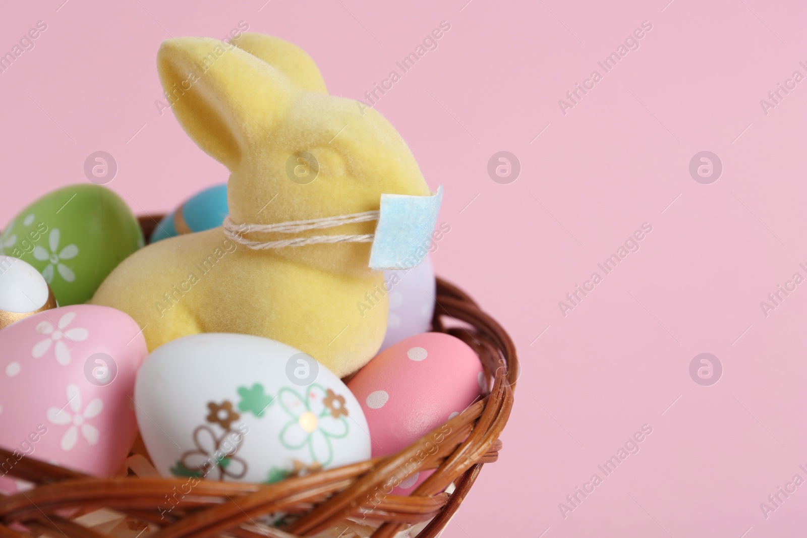 Photo of Cute bunny figure with protective mask and eggs in wicker basket on pink background, space for text. Easter holiday during COVID-19 quarantine