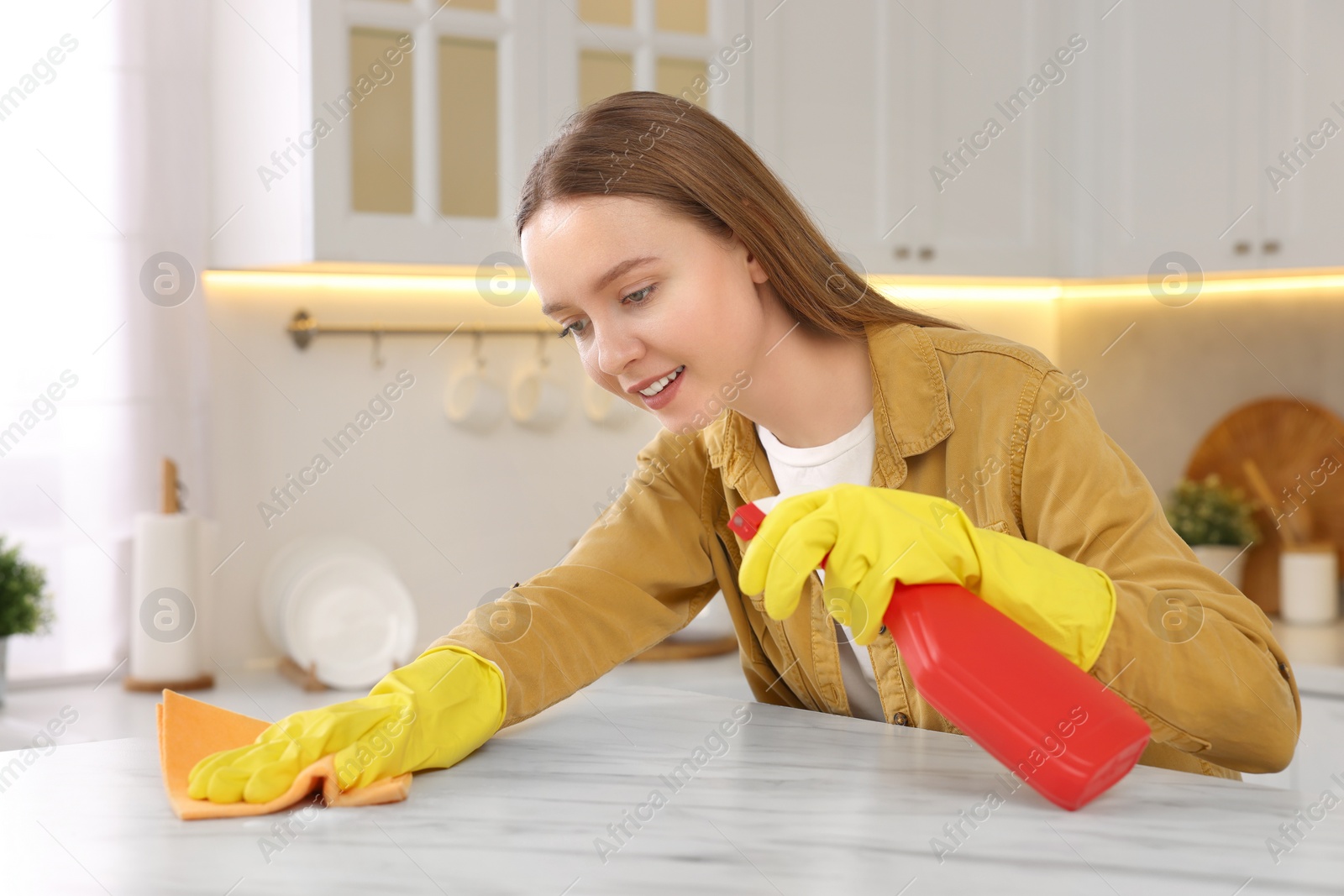 Photo of Woman with spray bottle and microfiber cloth cleaning white marble table in kitchen