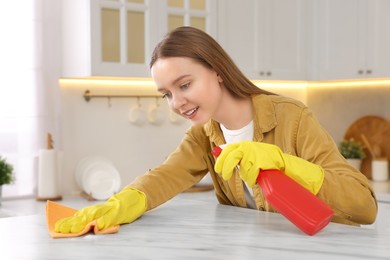 Photo of Woman with spray bottle and microfiber cloth cleaning white marble table in kitchen