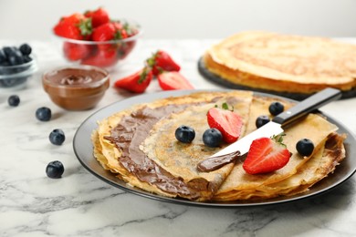 Photo of Delicious thin pancakes with chocolate paste and berries on white marble table