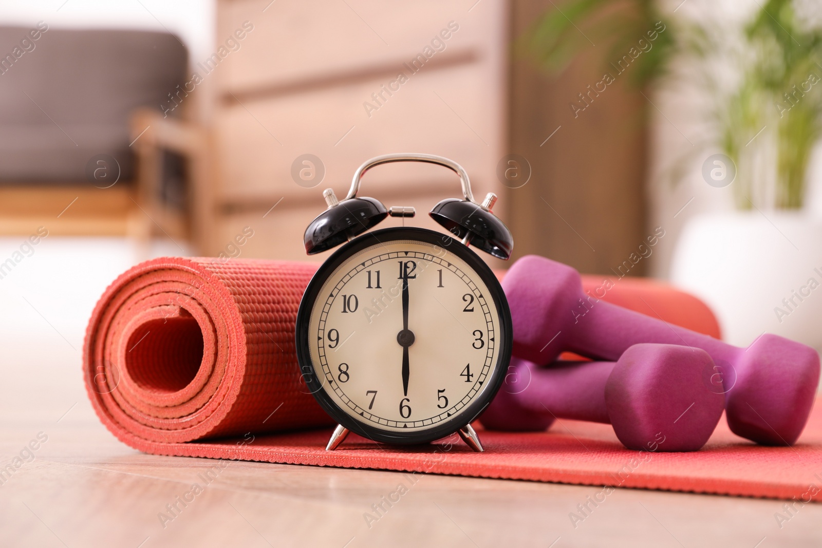 Photo of Alarm clock, yoga mat and dumbbells on wooden floor indoors. Morning exercise