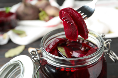 Fork with pickled beets over glass jar on wooden table, closeup