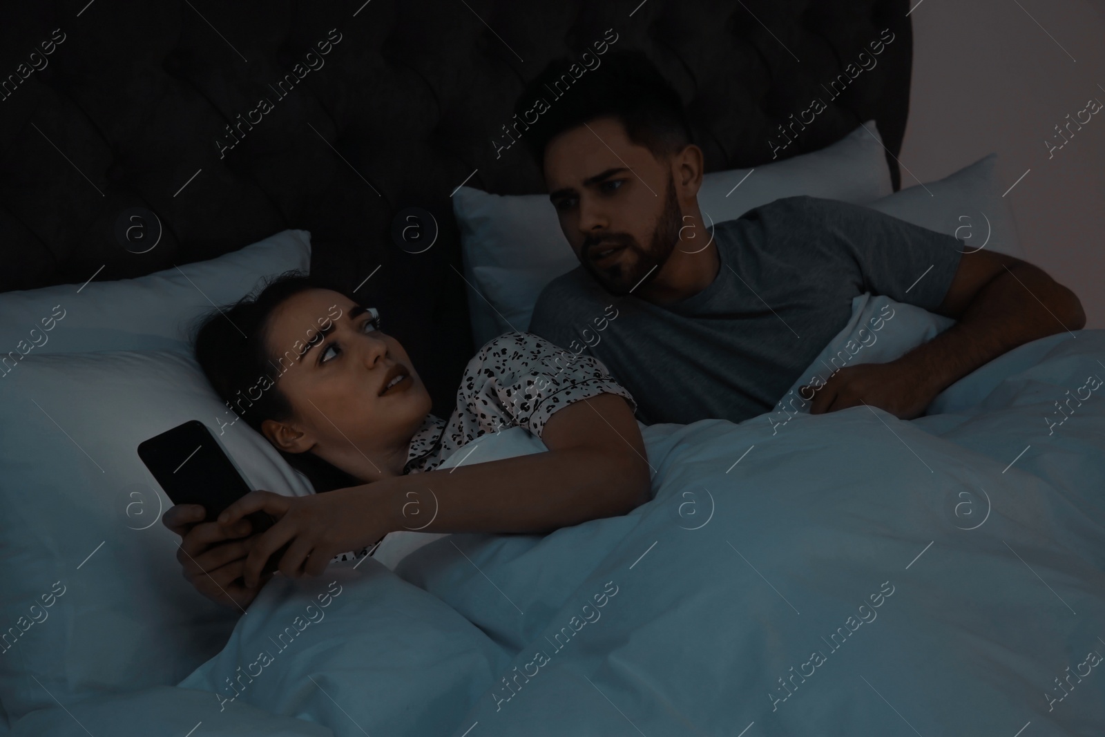 Photo of Distrustful young man peering into girlfriend's smartphone in bed at night