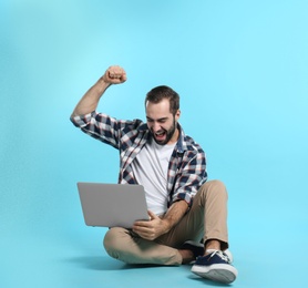 Emotional young man with laptop celebrating victory on color background