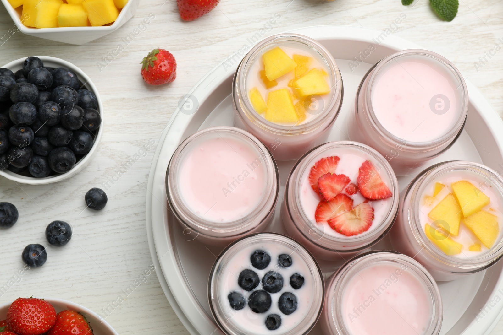 Photo of Yogurt maker with jars and different fruits on white wooden table, flat lay