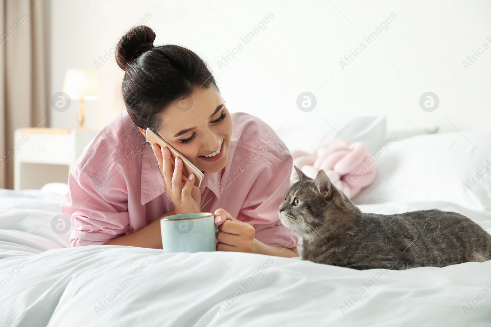 Photo of Young woman with cup of coffee talking on phone while lying near cute cat in bedroom. Pet and owner