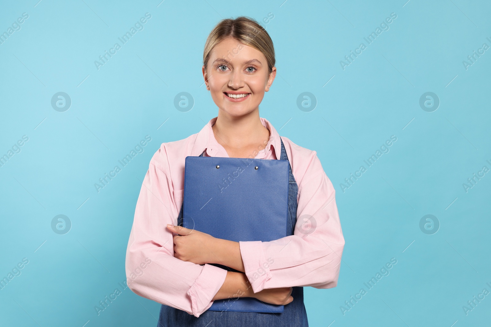 Photo of Beautiful young woman in denim apron with clipboard on light blue background