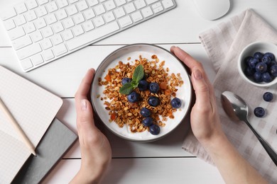 Photo of Woman holding bowl of tasty granola with blueberries at white wooden table with computer keyboard, top view