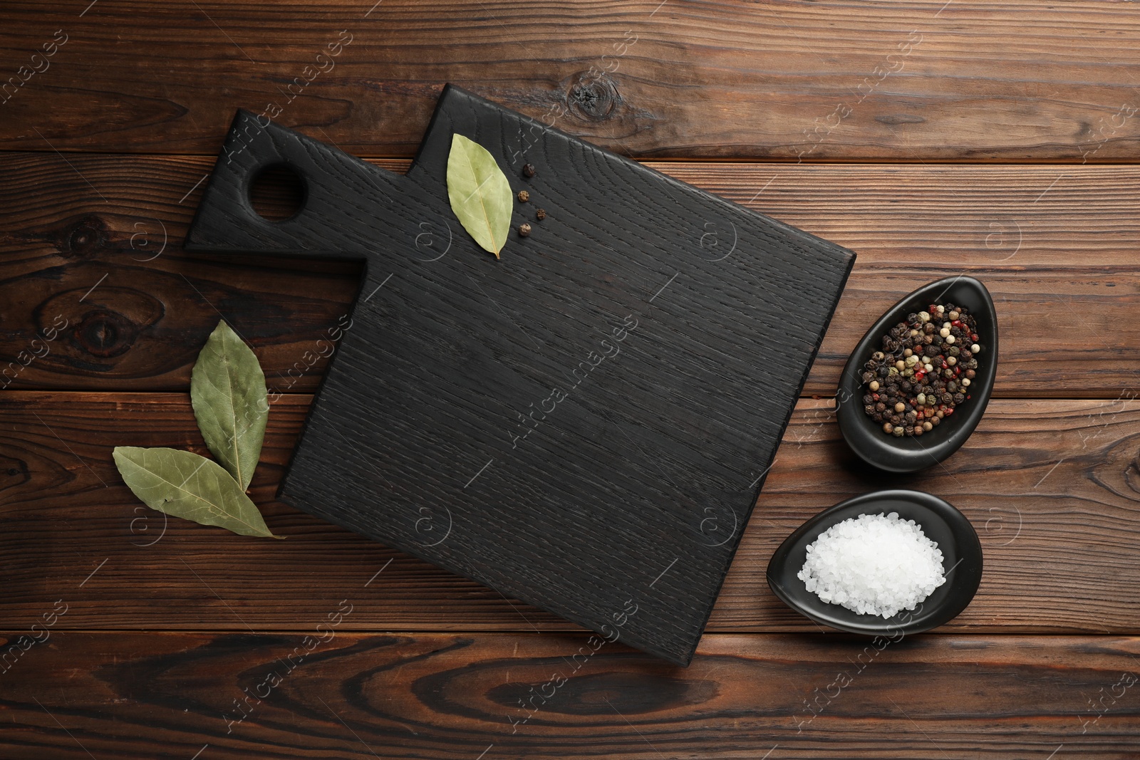 Photo of Black cutting board, salt, pepper and bay leaves on wooden table, flat lay. Space for text