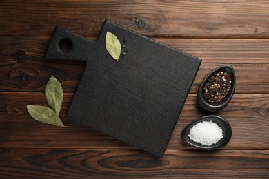 Photo of Black cutting board, salt, pepper and bay leaves on wooden table, flat lay. Space for text