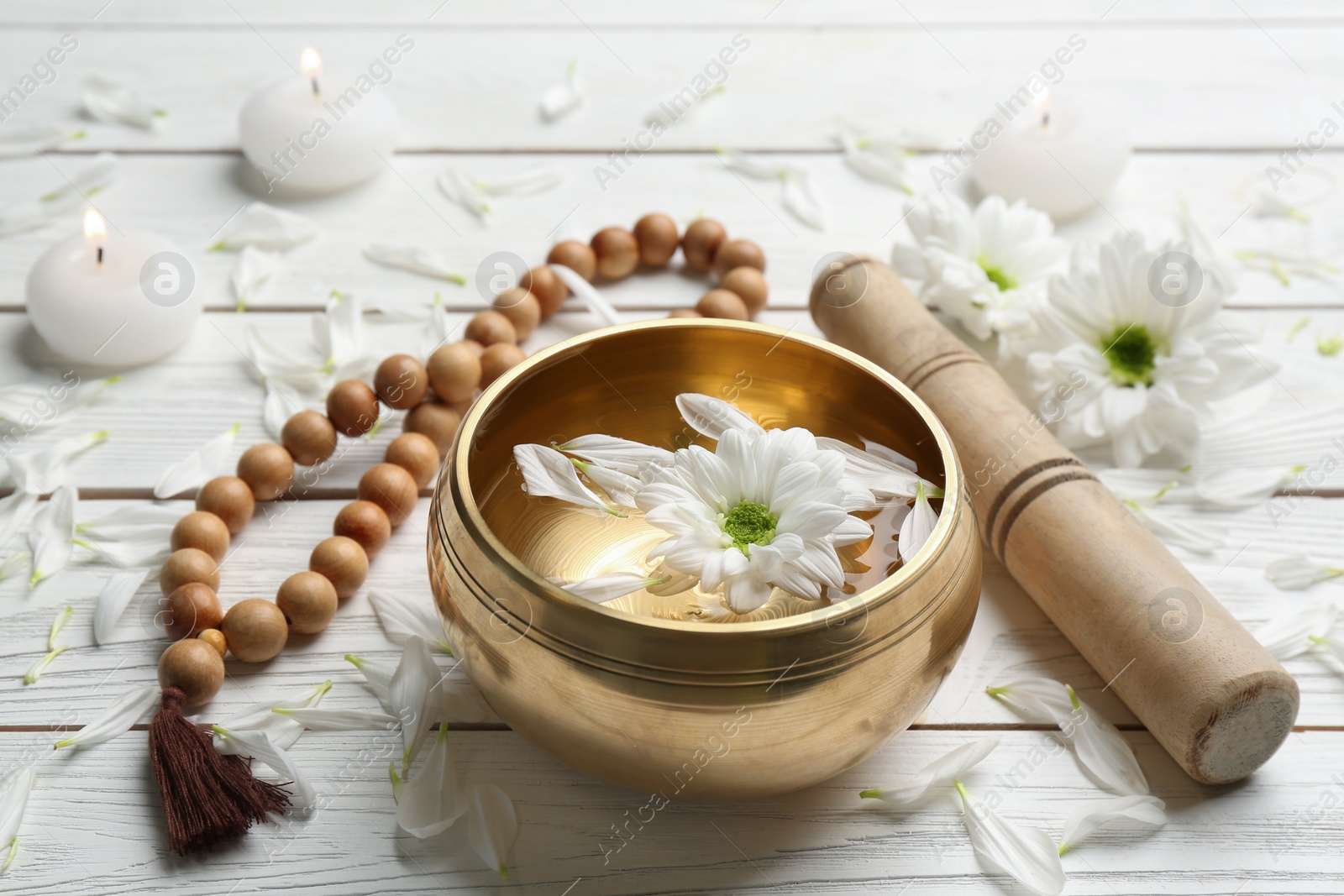 Photo of Golden singing bowl with flower, mallet and beads on white wooden table. Sound healing