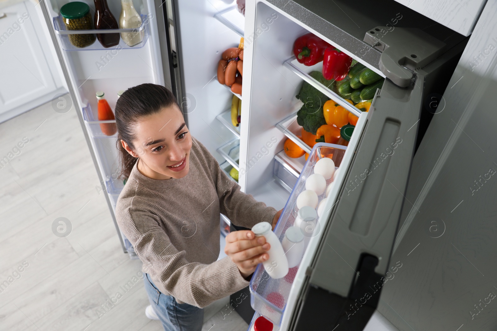 Photo of Young woman taking yoghurt out of refrigerator indoors, above view