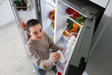 Photo of Young woman taking yoghurt out of refrigerator indoors, above view