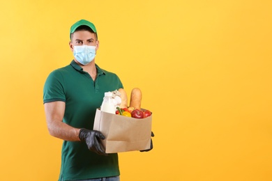 Courier in medical mask holding paper bag with food on yellow background, space for text. Delivery service during quarantine due to Covid-19 outbreak