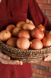 Woman holding wicker basket with ripe onions against red brick wall, closeup