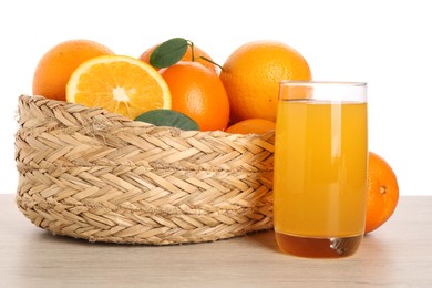 Fresh oranges in wicker basket and glass of juice on light wooden table against white background