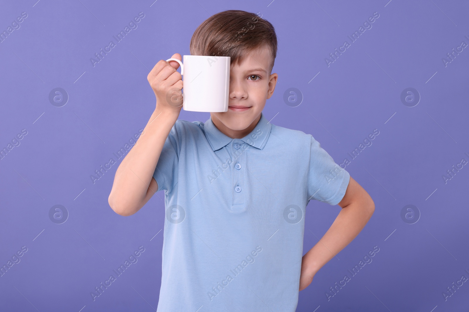 Photo of Cute boy covering eye with white ceramic mug on violet background