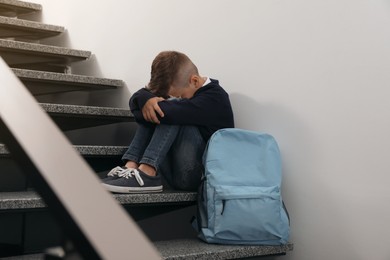 Upset little boy with backpack sitting on stairs indoors