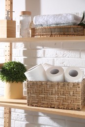 Photo of Toilet paper rolls in wicker basket and floral decor on wooden shelf near white brick wall