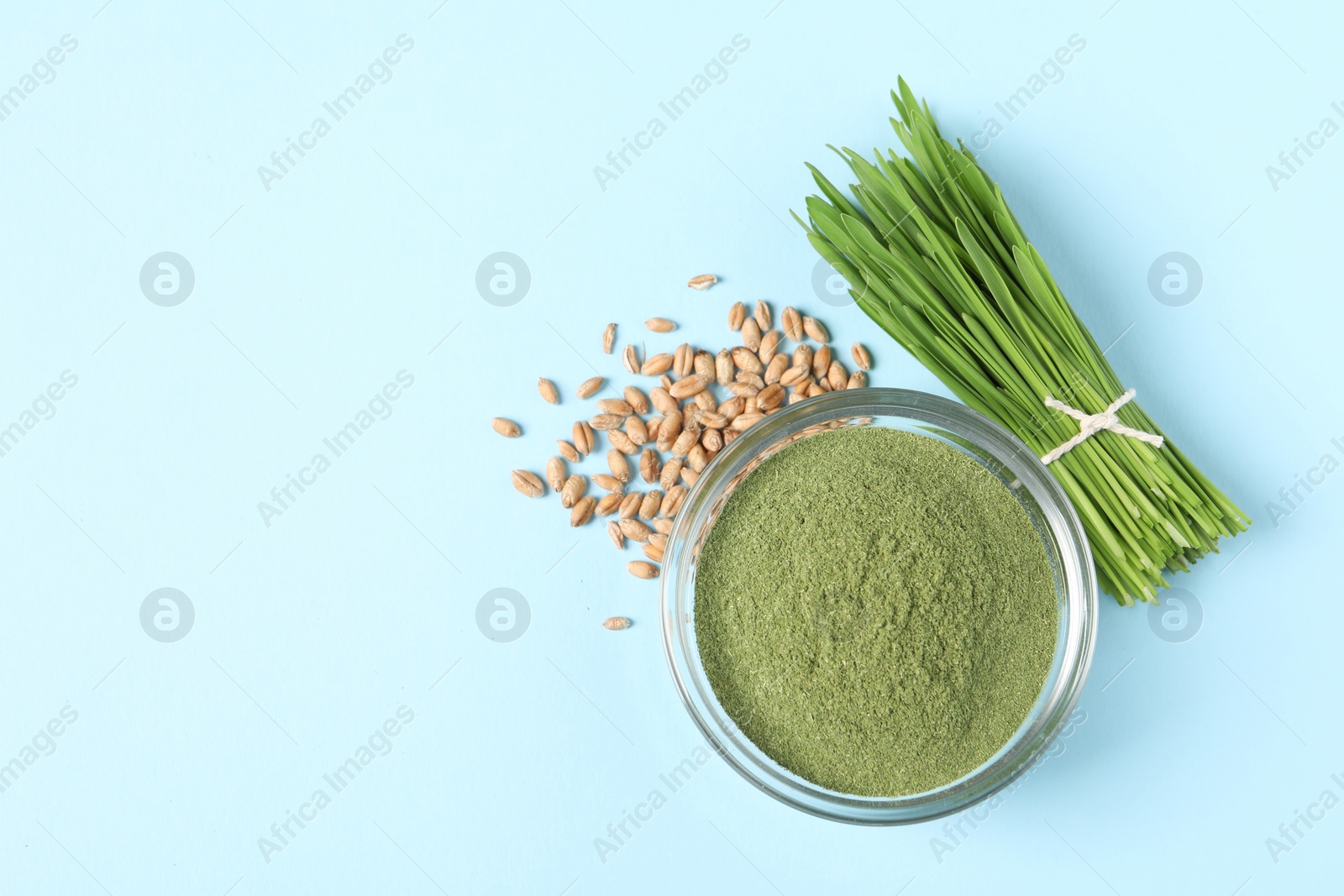 Photo of Wheat grass powder in glass bowl, seeds and fresh sprouts on light blue table, flat lay. Space for text