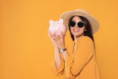 Photo of Young woman in stylish sunglasses and straw hat with piggy bank on orange background, space for text