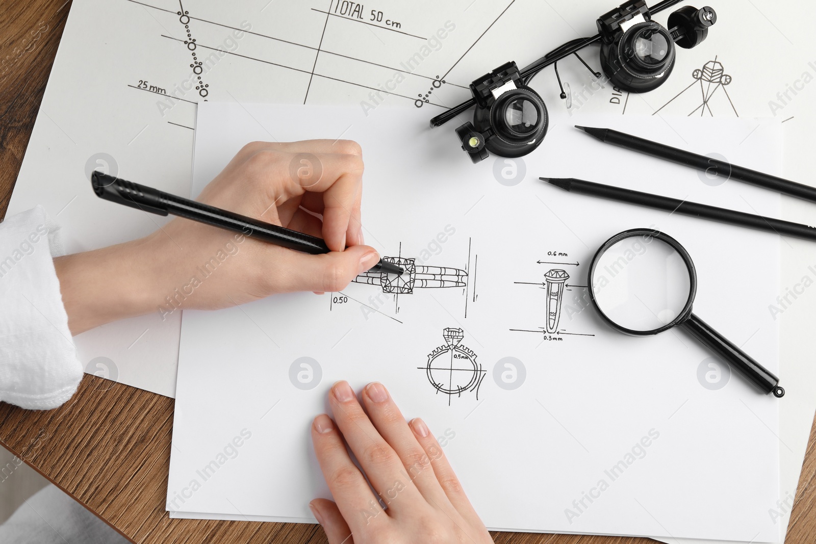 Photo of Jeweler drawing sketch of elegant ring on paper at wooden table, top view