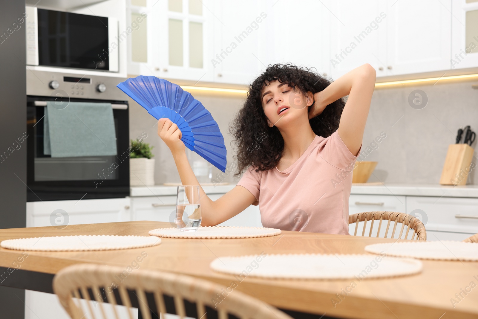 Photo of Young woman waving blue hand fan to cool herself at table in kitchen