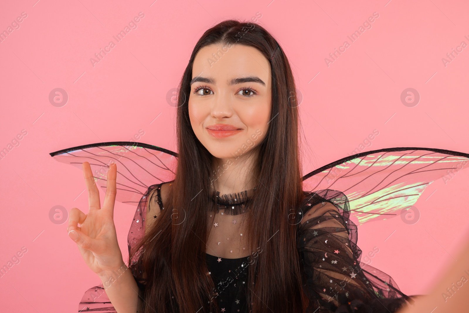 Photo of Beautiful girl in fairy costume with wings taking selfie and showing peace sign on pink background