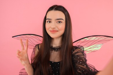 Beautiful girl in fairy costume with wings taking selfie and showing peace sign on pink background