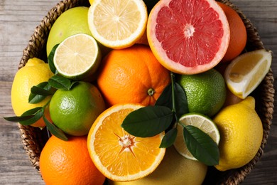 Photo of Different fresh citrus fruits and leaves in wicker basket on wooden table, top view