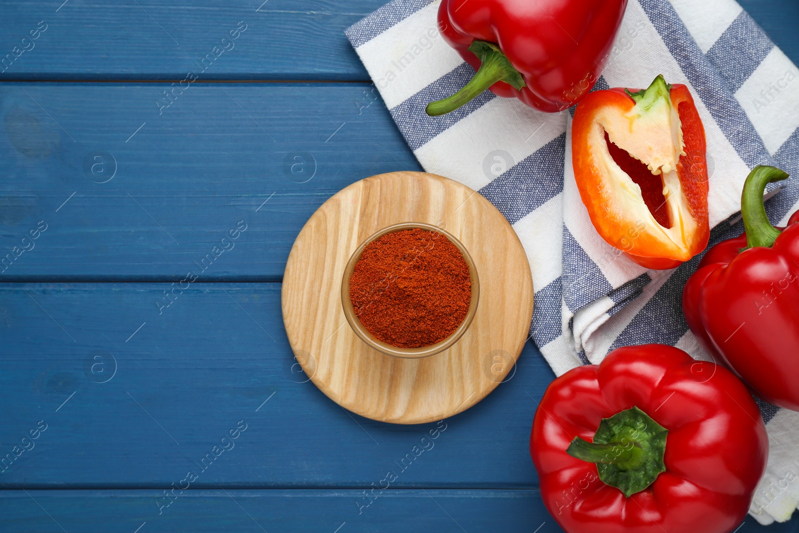 Photo of Bowl with aromatic paprika powder and fresh bell peppers on blue wooden table, flat lay. Space for text