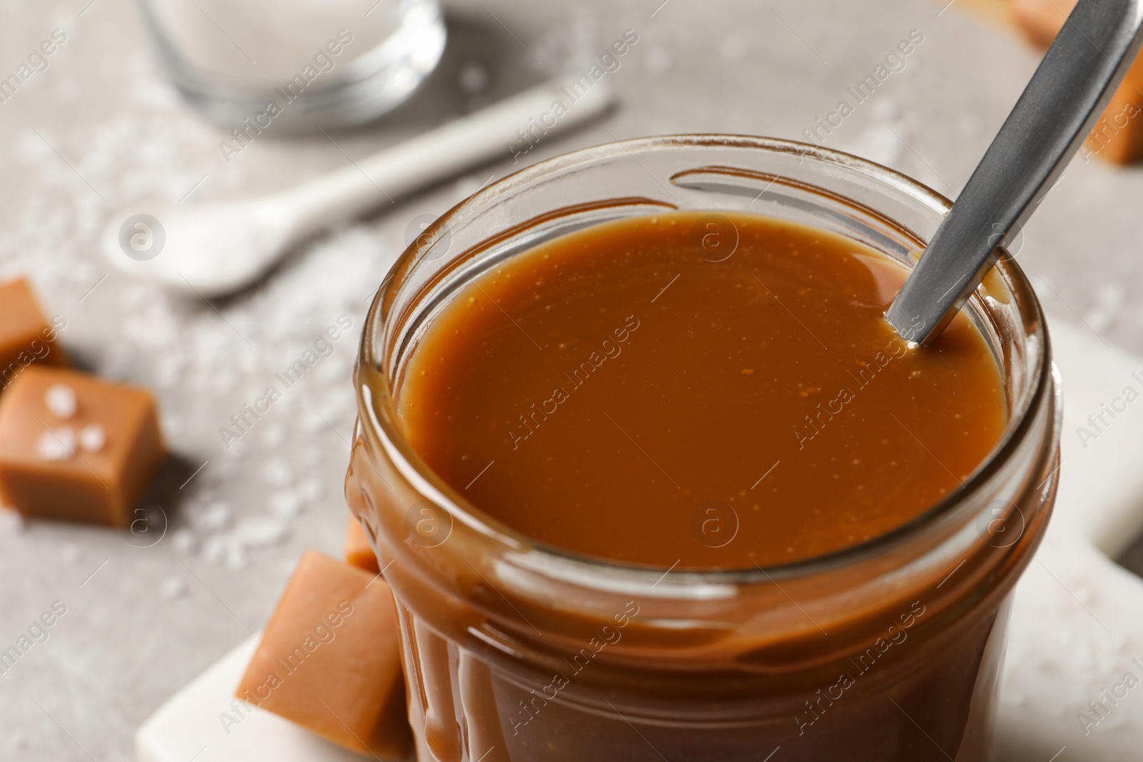 Photo of Tasty salted caramel and spoon in glass jar on table, closeup