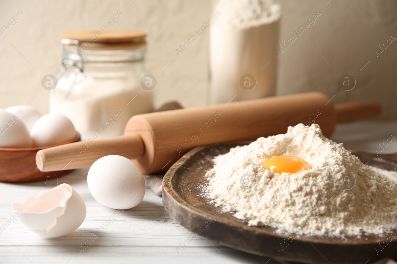 Photo of Making dough. Pile of flour with yolk, rolling pin and eggs on white wooden table, closeup