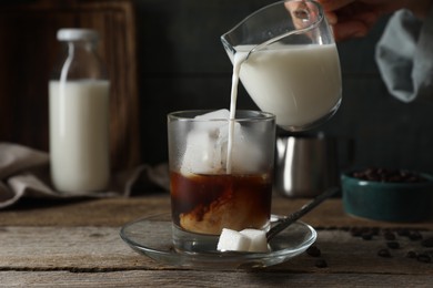 Woman pouring milk into glass with iced coffee at wooden table, closeup