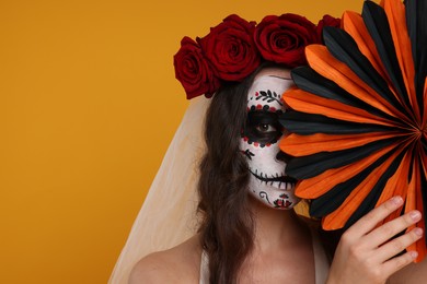 Photo of Young woman in scary bride costume with sugar skull makeup, flower crown and paper decoration on orange background, space for text. Halloween celebration
